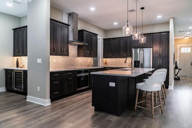 kitchen featuring appliances with stainless steel finishes, an island with sink, sink, hanging light fixtures, and wall chimney exhaust hood