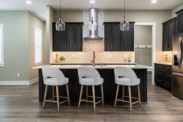 kitchen with a kitchen island with sink, hanging light fixtures, stainless steel refrigerator with ice dispenser, tasteful backsplash, and wall chimney exhaust hood