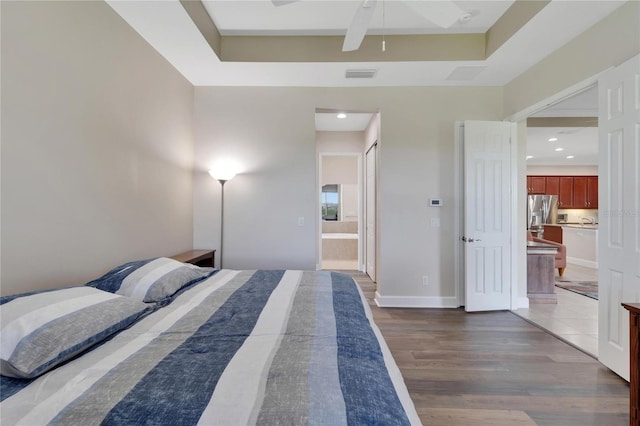 bedroom featuring stainless steel fridge, ceiling fan, light wood-type flooring, ensuite bath, and a raised ceiling