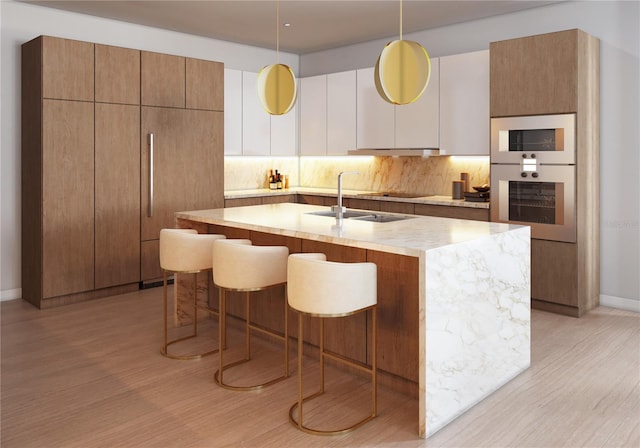 kitchen featuring wall oven, hanging light fixtures, decorative backsplash, an island with sink, and light hardwood / wood-style flooring