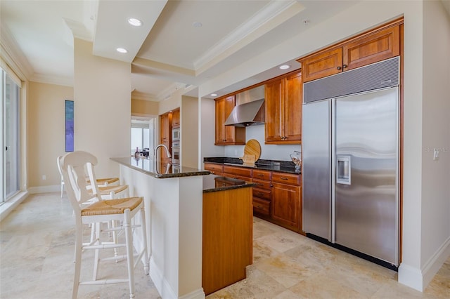 kitchen featuring a kitchen island with sink, wall chimney range hood, stainless steel appliances, dark stone countertops, and ornamental molding