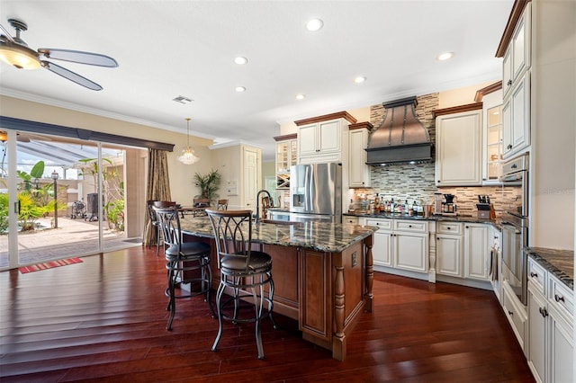 kitchen featuring dark wood-type flooring, stainless steel appliances, premium range hood, a kitchen island with sink, and backsplash