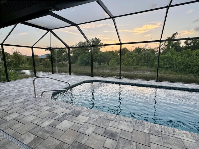 pool at dusk with glass enclosure, a water view, and a patio