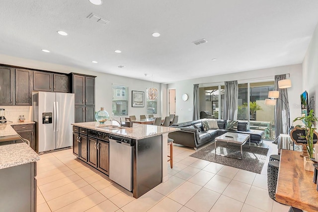 kitchen featuring a kitchen island with sink, light tile floors, dark brown cabinets, stainless steel appliances, and tasteful backsplash