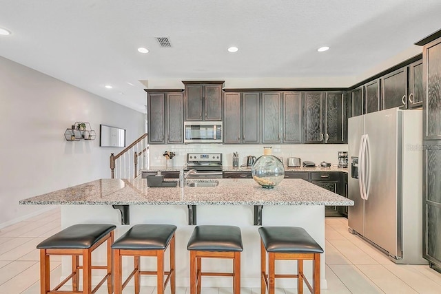 kitchen with a kitchen island with sink, appliances with stainless steel finishes, light stone countertops, and dark brown cabinetry