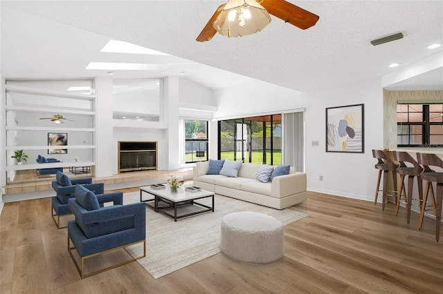 living room featuring ceiling fan, light wood-type flooring, a tiled fireplace, vaulted ceiling with skylight, and built in features