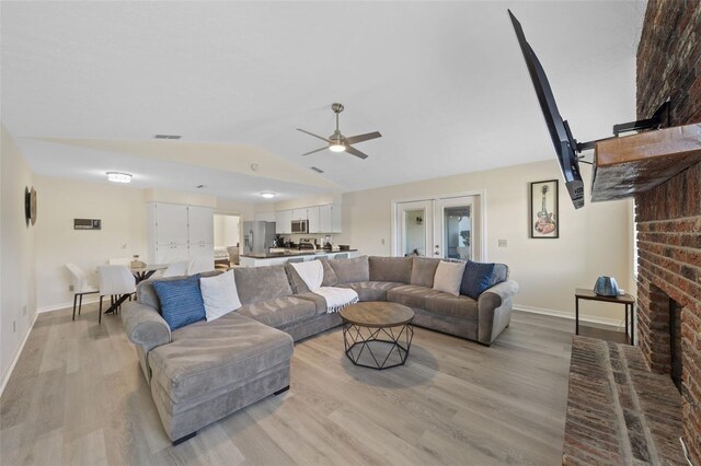 living room featuring french doors, a brick fireplace, vaulted ceiling, ceiling fan, and light wood-type flooring