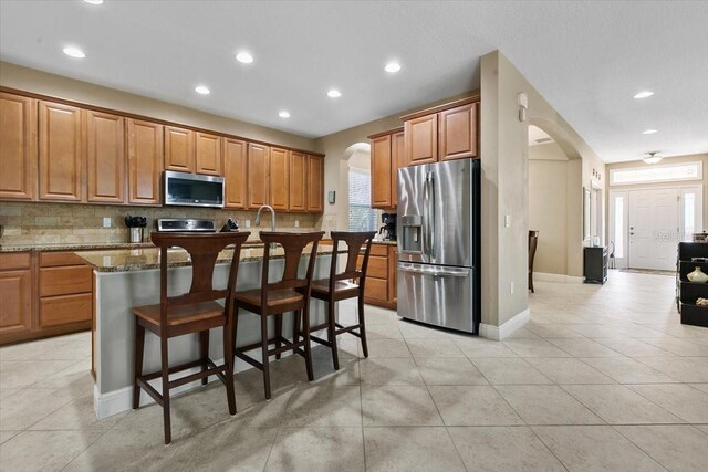 kitchen with light stone counters, stainless steel appliances, an island with sink, light tile patterned floors, and a breakfast bar area