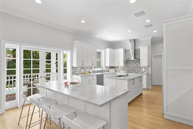 kitchen with light wood-type flooring, a center island with sink, wall chimney range hood, and white cabinetry