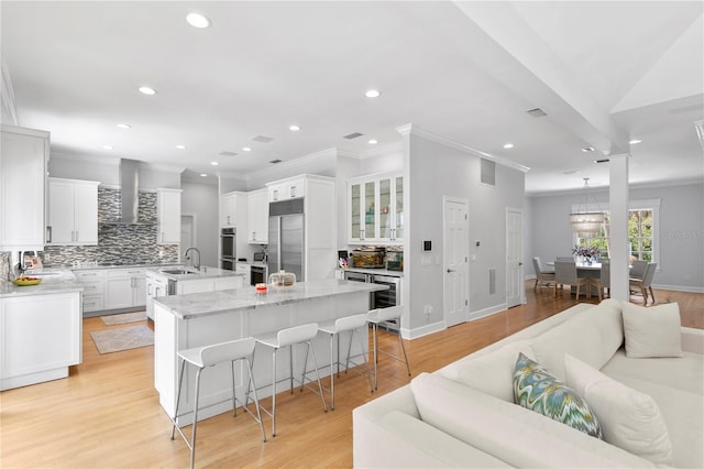 kitchen featuring white cabinets, wall chimney range hood, stainless steel appliances, a breakfast bar area, and an island with sink