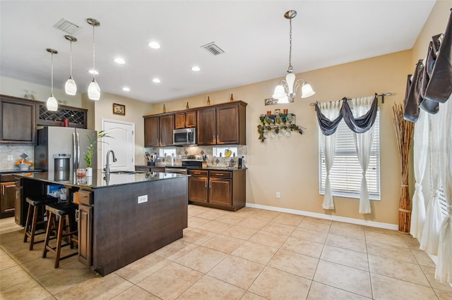 kitchen featuring light tile floors, stainless steel appliances, an island with sink, dark brown cabinets, and hanging light fixtures