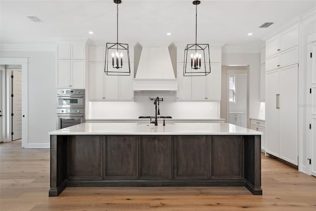 kitchen with double oven, decorative light fixtures, an island with sink, and premium range hood