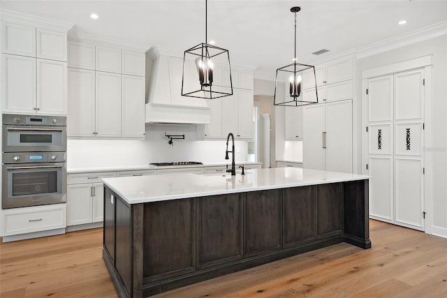 kitchen with premium range hood, white cabinets, ornamental molding, a kitchen island with sink, and stainless steel appliances