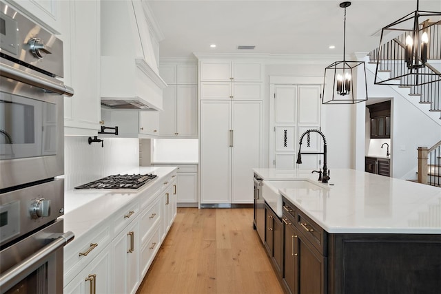kitchen featuring premium range hood, decorative light fixtures, white cabinetry, sink, and a center island with sink