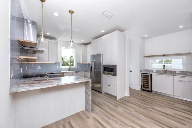 kitchen with appliances with stainless steel finishes, wall chimney range hood, decorative light fixtures, light hardwood / wood-style flooring, and white cabinets