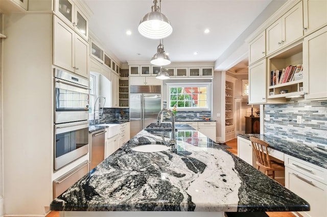 kitchen featuring built in desk, dark stone countertops, a large island with sink, hanging light fixtures, and stainless steel appliances