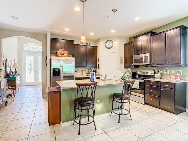 kitchen featuring light stone countertops, a kitchen bar, appliances with stainless steel finishes, an island with sink, and pendant lighting