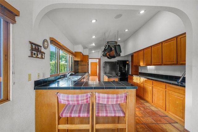 kitchen with tile countertops, a kitchen bar, kitchen peninsula, black refrigerator, and sink