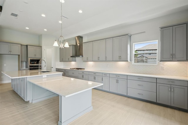 kitchen with a center island with sink, wall chimney exhaust hood, appliances with stainless steel finishes, gray cabinetry, and a sink