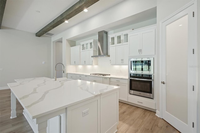 kitchen featuring a center island with sink, beamed ceiling, stainless steel appliances, and wall chimney range hood