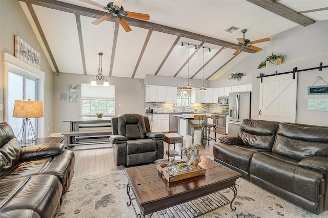 living room featuring ceiling fan, light hardwood / wood-style flooring, beam ceiling, a barn door, and high vaulted ceiling