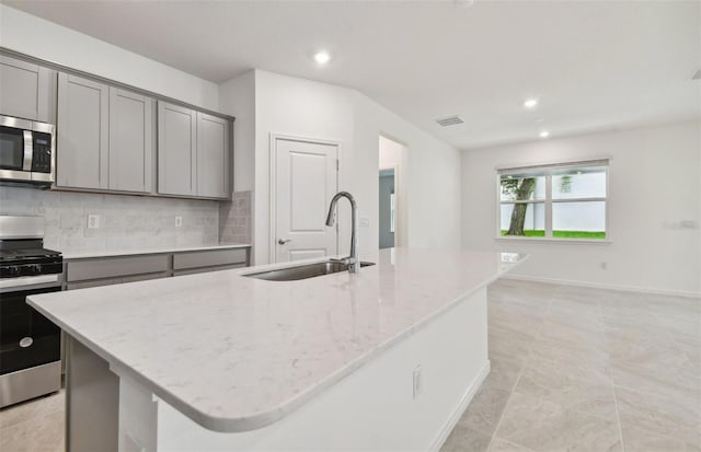 kitchen featuring sink, gray cabinetry, light stone counters, appliances with stainless steel finishes, and a kitchen island with sink