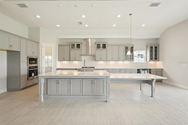 kitchen featuring wall chimney exhaust hood, an island with sink, stainless steel microwave, gray cabinetry, and hanging light fixtures