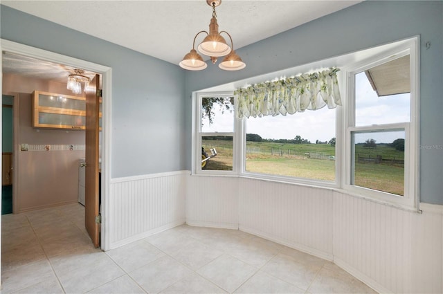 unfurnished dining area with a wainscoted wall, light tile patterned floors, a notable chandelier, and a rural view