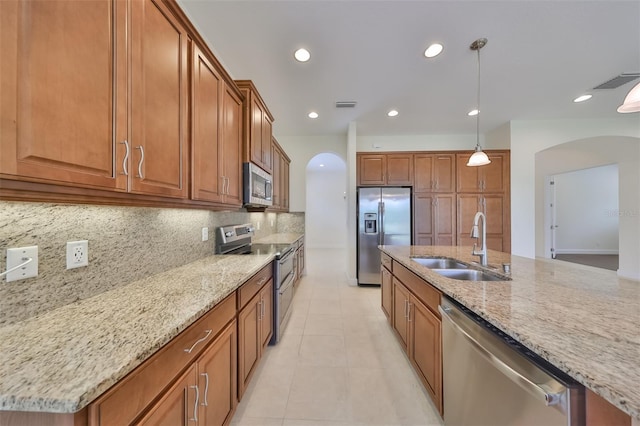 kitchen with light stone counters, stainless steel appliances, a kitchen island with sink, sink, and pendant lighting