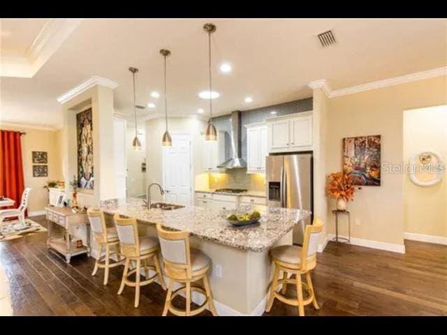 kitchen with hanging light fixtures, wall chimney range hood, a breakfast bar, stainless steel fridge, and white cabinetry