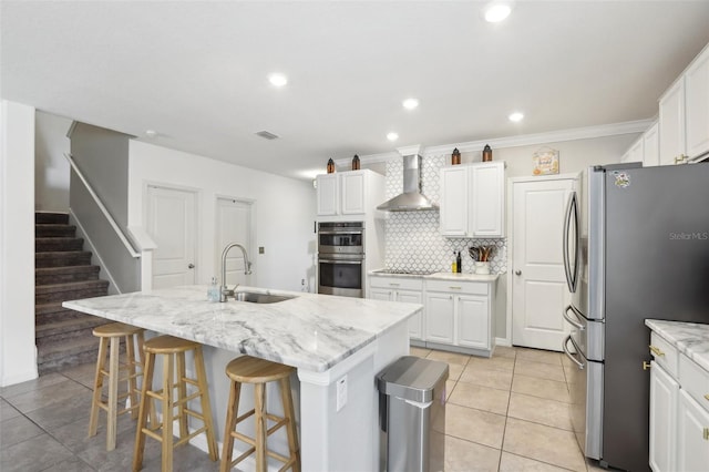 kitchen featuring stainless steel appliances, a sink, white cabinets, wall chimney range hood, and an island with sink