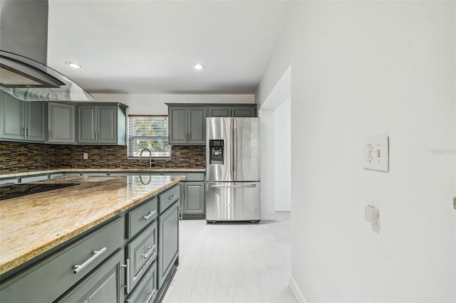 kitchen featuring decorative backsplash, stainless steel refrigerator with ice dispenser, light stone countertops, island exhaust hood, and sink