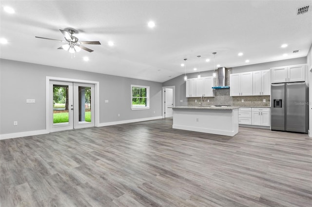 kitchen with pendant lighting, stainless steel fridge, white cabinetry, a kitchen island with sink, and wall chimney exhaust hood