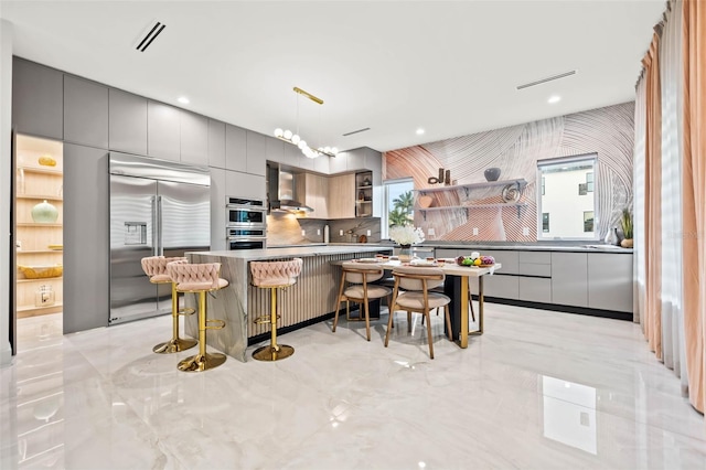 kitchen featuring gray cabinetry, stainless steel appliances, wall chimney exhaust hood, and hanging light fixtures