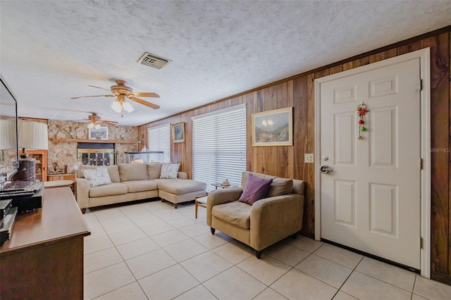 living room with ceiling fan, light tile patterned flooring, wood walls, a stone fireplace, and a textured ceiling