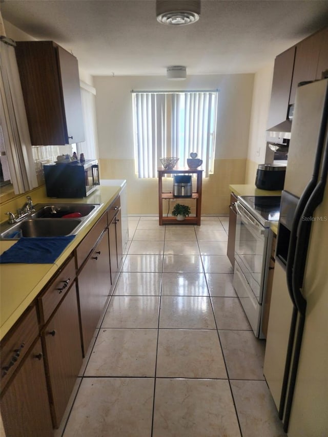 kitchen with wall chimney range hood, dark brown cabinetry, appliances with stainless steel finishes, sink, and light tile floors