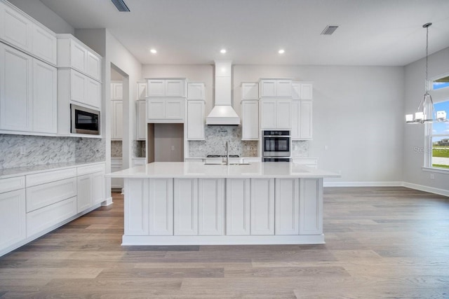 kitchen with light wood-style flooring, premium range hood, built in microwave, tasteful backsplash, and a large island with sink