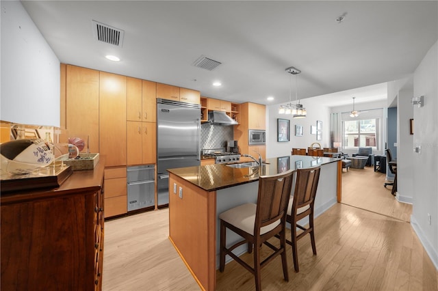 kitchen with built in appliances, an island with sink, pendant lighting, light hardwood / wood-style floors, and decorative backsplash