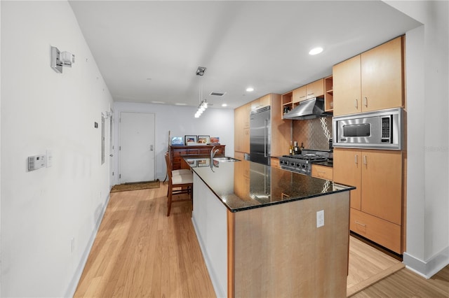 kitchen with hanging light fixtures, light hardwood / wood-style flooring, built in appliances, an island with sink, and decorative backsplash