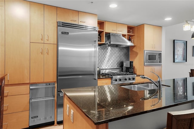 kitchen with wall chimney exhaust hood, tasteful backsplash, built in appliances, dark stone countertops, and a kitchen island with sink