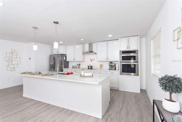 kitchen with a kitchen island with sink, white cabinets, wall chimney range hood, light wood-type flooring, and stainless steel appliances