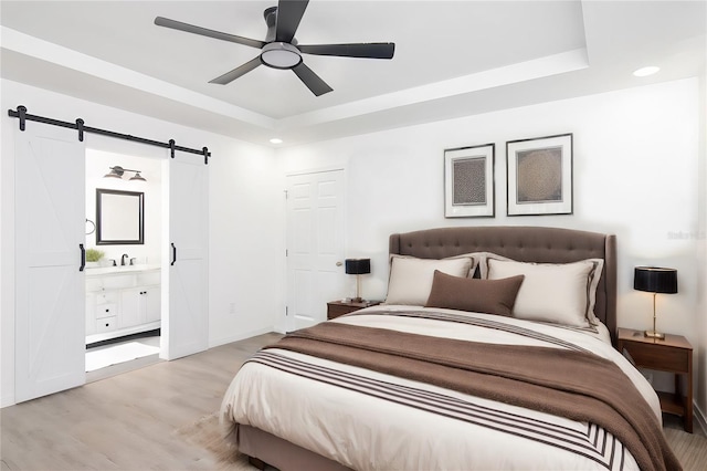 bedroom featuring ensuite bathroom, a raised ceiling, ceiling fan, a barn door, and light hardwood / wood-style floors