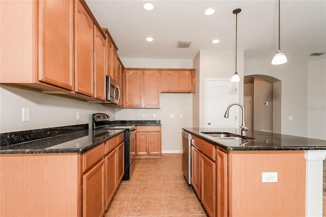 kitchen featuring dark stone counters, stainless steel appliances, sink, decorative light fixtures, and an island with sink