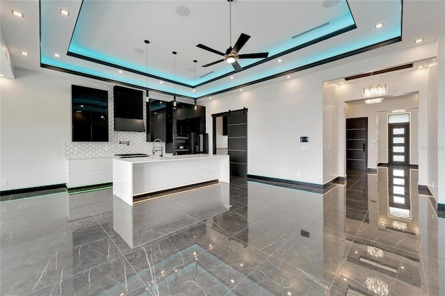 kitchen with ceiling fan, a barn door, an island with sink, and a tray ceiling