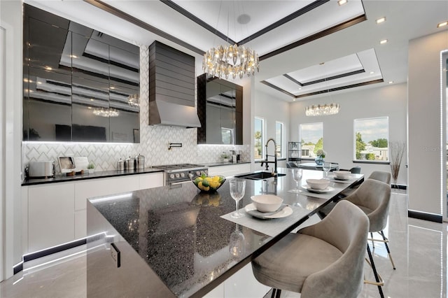 kitchen with backsplash, a raised ceiling, a kitchen island with sink, and hanging light fixtures