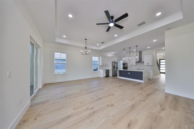 unfurnished living room with light wood-type flooring, ceiling fan with notable chandelier, and a raised ceiling