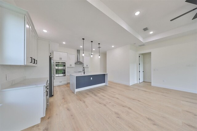 kitchen with ceiling fan, decorative light fixtures, a kitchen island with sink, wall chimney exhaust hood, and white cabinetry