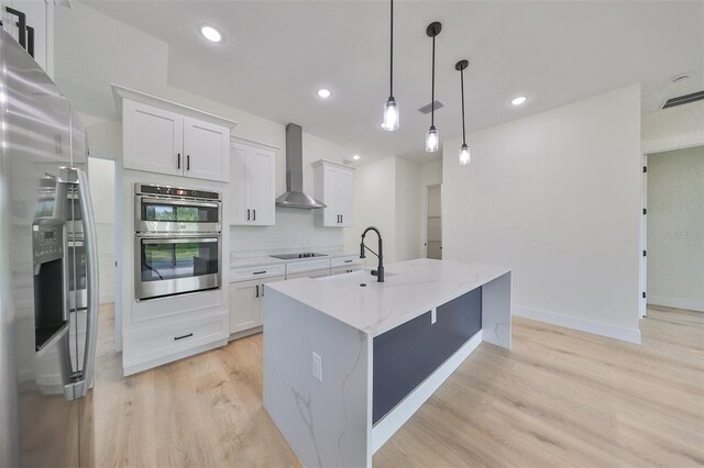 kitchen featuring white cabinets, stainless steel appliances, decorative light fixtures, a kitchen island with sink, and wall chimney range hood