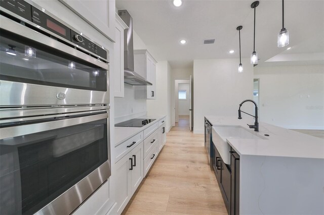 kitchen featuring appliances with stainless steel finishes, light hardwood / wood-style floors, white cabinets, wall chimney exhaust hood, and decorative light fixtures