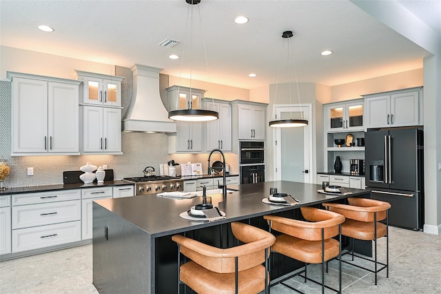 kitchen featuring an island with sink, decorative light fixtures, black appliances, premium range hood, and light tile floors